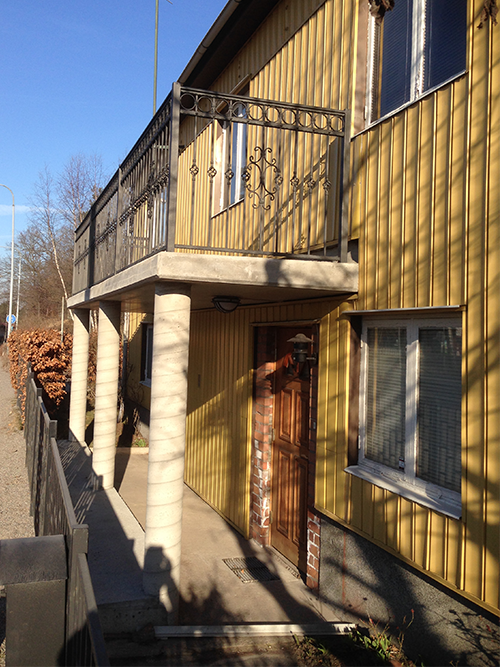 Platsgjuten balkong med runda pelare och smidesräcke på gul villa.
